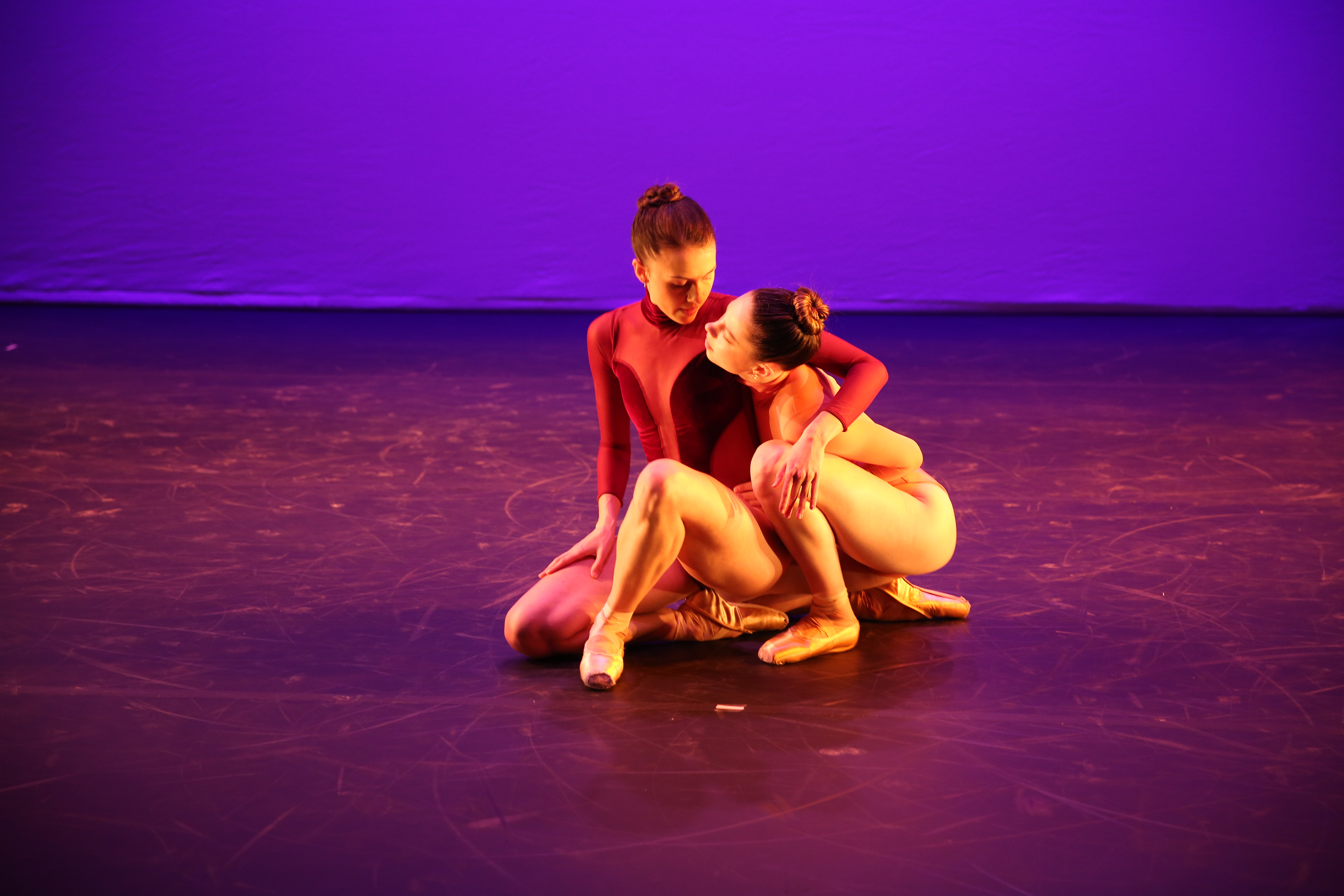 Remy Young and Sierra Armstrong are seated and embracing each other on stage. The stage background is purple the women have their hair in tight buns and wear leotards, no tights, and pointe shoes. Remy's leotard is light red with darker red details, we can't see Sierra's outfit very well. 